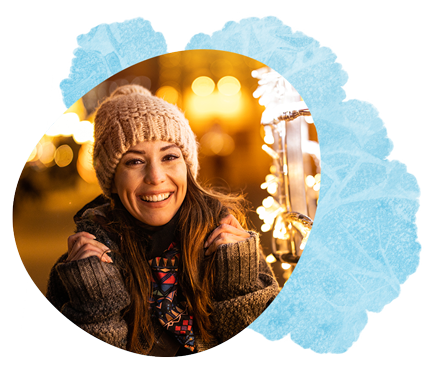 Smiling woman wearing a knit hat and scarf outdoors in a festive winter setting.